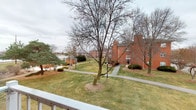 This single-floor unit offers a thoughtfully arranged layout with comfortable, carpeted living spaces and several notable features throughout. The living room serves as the heart of the home, anchored by a classic brick fireplace with a white-painted mantle surround and glass-front doors — a standout architectural detail that adds warmth and character to the space. The room is bright and open, with white walls and neutral carpet that create a clean, versatile canvas.

A glass balcony door opens to a private outdoor deck, framed by a decorative wrought iron railing. The balcony overlooks a well-maintained courtyard featuring mature trees, manicured lawns, walking paths, and charming lamp posts, with views extending toward a neighboring brick building and what appears to be a nearby waterway — providing a peaceful, park-like setting.

The interior continues with a hallway connecting the living area to additional rooms, including a kitchen and closeted storage spaces. Interior doors are classic six-panel white, consistent throughout the unit. The overall ambiance is clean and neutral, offering a welcoming and move-in-ready environment. The combination of the cozy fireplace, private balcony, and serene outdoor views makes this a distinctive and comfortable space.