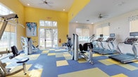 This fitness center is a vibrant, well-appointed space designed to support a full range of workout activities. The interior is immediately eye-catching, featuring bold yellow accent walls paired with crisp white ceilings and a striking blue, yellow, and green checkered tile floor that adds energy and personality to the environment. Recessed lighting and multiple ceiling fans ensure the space remains bright and comfortable throughout.

The facility is thoughtfully divided into distinct exercise zones. The cardio area is equipped with a row of treadmills and stationary bikes, each with individual monitor screens, positioned near large windows fitted with roll-up shades that invite natural light and views of the surrounding outdoor greenery. French doors along one wall provide additional light and direct outdoor access.

The strength training section houses a comprehensive selection of equipment, including a Hoist cable machine, a power rack with a bench, and a dumbbell rack stocked with a range of free weights. Full-length mirrors along one wall allow users to monitor their form during exercise.

A dedicated area toward the rear of the space provides room for additional machines and cycling equipment. Wall-mounted informational boards and a fire safety station reflect the facility's attention to safety and user awareness. The overall ambiance is bright, motivating, and well-organized, making this fitness center a fully functional and inviting space for exercise.