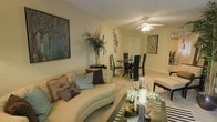 This single-floor residence offers an inviting open-concept layout that effortlessly blends the living and dining areas into one cohesive, functional space. The living area is anchored by a generously sized curved sofa adorned with an assortment of decorative pillows, complemented by a sleek glass coffee table that adds a contemporary touch. Warm, neutral tones on the walls create a calm and welcoming ambiance throughout. Decorative wall art, including a striking tree silhouette canvas and botanical accents, adds visual interest and personality to the space. A tall indoor palm plant introduces a natural, organic element that enhances the overall atmosphere. The dining area, positioned just beyond the living room, features a glass-top dining table surrounded by elegant dark upholstered chairs, making it ideal for both casual meals and entertaining guests. Overhead, a ceiling fan with a light fixture provides both comfort and illumination, contributing to the space's airy feel. Light-toned carpeting runs throughout, tying the two areas together harmoniously. The thoughtful arrangement of furniture and decor reflects a refined, contemporary aesthetic that is both practical and stylish.