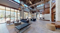 This impressive single-story space showcases a masterful blend of rustic and contemporary design elements throughout its generously proportioned layout. The centerpiece is a grand open lounge area distinguished by dramatically soaring ceilings supported by substantial exposed wood beams and natural limestone columns, creating a warm yet refined architectural statement. Large floor-to-ceiling steel-framed windows line the perimeter, drawing in abundant natural light and offering views of an outdoor terrace and pool area beyond.

The main lounge is thoughtfully furnished with multiple seating arrangements, including tufted leather benches, navy blue sofas, and sleek accent chairs positioned atop layered area rugs over large-format stone tile flooring. Wrought iron chandelier pendants hang from the beam structure above, complementing the wall-mounted sconces throughout. A stone-clad fireplace anchors one wall, while a bar area with counter seating provides an additional gathering space.

Adjacent areas include a cozy reading nook with leather armchairs, studded ottomans, and a glass-front credenza flanked by tall floor lamps. A well-appointed kitchenette features rich wood cabinetry, light stone countertops, and stainless steel appliances set against the recurring natural limestone walls. A fitness room is also accessible from the main corridor, completing this versatile and elegantly designed communal facility.