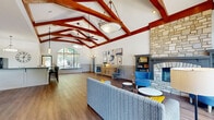 This single-floor clubhouse in Cordova, Tennessee, offers a thoughtfully designed communal space defined by striking architectural details and a warm, welcoming ambiance. The centerpiece of the main living area is a soaring vaulted ceiling adorned with bold, rich-toned wood beams that draw the eye upward, creating a sense of grandeur throughout the open floor plan. Wide-plank hardwood flooring flows seamlessly across the entire space, complementing the neutral wall tones accented by classic wainscoting.

The living area features a floor-to-ceiling stacked stone fireplace flanked by built-in shelving, providing a cozy focal point. Large arched windows flood the space with abundant natural light and offer views of the surrounding greenery. Comfortable seating arrangements invite relaxation and social gathering.

The full kitchen is equipped with a generous island bar featuring dark countertops, bar stool seating, pendant lighting, and a full-size refrigerator — ideal for catering events and community functions. An adjacent dining area with a round table and modern pendant fixture provides a casual space for meals and meetings. A dedicated theater room, a bathroom, and additional flex spaces round out the layout, making this clubhouse a versatile and functional hub for community life.