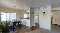 This single-floor residence is designed with an open-concept layout that seamlessly connects the dining, kitchen, and living areas, creating a spacious and functional flow. The interior is finished with warm hardwood-style flooring in the main living and dining spaces, transitioning to plush carpet in the living area, offering a comfortable contrast underfoot.

The dining area is defined by a stylish pendant light fixture that hangs above a bar-height table with matching stools, adding a contemporary touch. A sunburst clock decorates the wall, complementing the eclectic décor accents, including antler wall art and a framed feather print.

The kitchen is partially separated by a half-wall with a pass-through counter, providing both visual openness and practical division. White cabinetry, a white refrigerator, and a window above the sink allow natural light to brighten the space. Overhead lighting keeps the kitchen well-illuminated.

The neutral, light-toned walls throughout create a clean and airy ambiance, while HVAC vents in the ceiling suggest a centralized climate control system. The overall design balances an inviting, casual aesthetic with practical functionality.