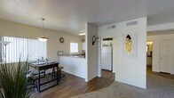 This single-floor residence is designed with an open-concept layout that seamlessly connects the dining, kitchen, and living areas, creating a spacious and functional flow. The interior is finished with warm hardwood-style flooring in the main living and dining spaces, transitioning to plush carpet in the living area, offering a comfortable contrast underfoot.

The dining area is defined by a stylish pendant light fixture that hangs above a bar-height table with matching stools, adding a contemporary touch. A sunburst clock decorates the wall, complementing the eclectic décor accents, including antler wall art and a framed feather print.

The kitchen is partially separated by a half-wall with a pass-through counter, providing both visual openness and practical division. White cabinetry, a white refrigerator, and a window above the sink allow natural light to brighten the space. Overhead lighting keeps the kitchen well-illuminated.

The neutral, light-toned walls throughout create a clean and airy ambiance, while HVAC vents in the ceiling suggest a centralized climate control system. The overall design balances an inviting, casual aesthetic with practical functionality.