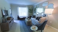 This single-floor space offers a thoughtfully designed and elegantly appointed interior that balances comfort with contemporary style. The living room serves as the heart of the space, showcasing crown molding along the ceilings and neutral-toned walls that create a calm, sophisticated atmosphere. Large windows flood the room with natural light, framing outdoor views and enhancing the airy feel of the interior.

The living area is furnished with a patterned sofa accented by plush decorative pillows, complemented by a sleek navy armchair and a light wood coffee table with open shelving below. A rich blue area rug anchors the seating arrangement, adding depth and warmth to the space. An arrangement of decorative mirrors in varying ornate shapes adorns one wall, adding visual interest and reflecting light throughout the room. A tall indoor palm plant introduces a touch of greenery, while a tufted bench positioned near the window provides an inviting reading nook.

The overall ambiance is polished yet welcoming, with carefully curated décor elements — from mirrored accents to layered textiles — that lend the space a refined, model-home quality. The layout feels open and well-proportioned, making it both functional and visually appealing.