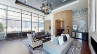This stylish single-floor space exudes a contemporary, upscale ambiance through a thoughtful combination of architectural details and curated design elements. The lounge area is anchored by a dramatic sputnik-style chandelier with metallic, sculptural arms that serves as an immediate focal point beneath a dark-painted ceiling, creating a bold contrast with the light-toned walls.

Floor-to-ceiling glass windows line one side of the space, flooding the interior with natural light while offering commanding views of a surrounding urban skyline. The dark hardwood flooring adds warmth and grounding throughout the open layout.

Seating arrangements feature a large, plush sectional adorned with an array of white and charcoal accent pillows, complemented by tufted armchairs positioned atop a geometric-patterned area rug. A curved wood-paneled architectural element creates a semi-private nook in the background, complete with built-in shelving and intimate booth-style seating — adding visual depth and a sense of layered zones within the space.

A textured stone accent wall and a built-in bar or refreshment station with a beverage refrigerator are also visible, rounding out the space's functional and aesthetic appeal.