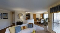 Located at 601 Jones Ferry Rd in Carrboro, NC, this single-floor residence offers a thoughtfully designed open-concept layout that seamlessly connects the living, dining, and kitchen areas. The living room serves as the heart of the home, featuring neutral wall tones accented by warm earth-toned décor, a plush seating area anchored by a patterned area rug, and a decorative round mirror that adds a stylish focal point. Natural light filters in through well-placed windows, lending a bright and welcoming atmosphere throughout. The kitchen is positioned just beyond the living area and features dark cabinetry, stainless steel appliances, and a peninsula-style counter that provides both workspace and a casual seating option. A bold accent wall in a rich golden hue defines the dining area, which is illuminated by an elegant pendant light fixture and complemented by a glass-top dining table with white upholstered chairs. Greenery and floral accents are incorporated throughout, contributing to a fresh, vibrant ambiance. The consistent use of wood-look flooring and coordinated finishes ties the entire space together, creating a cohesive and polished interior that is both functional and aesthetically refined.