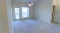 This single-floor space presents a clean, neutral aesthetic well-suited for comfortable everyday living. The main living area is finished with plush wall-to-wall carpeting in a light, neutral tone that complements the soft beige walls throughout. A ceiling fan with an integrated light fixture is centrally mounted overhead, providing both air circulation and ambient lighting to the room.

Natural light is a defining feature of this space, with a large window fitted with horizontal blinds allowing adjustable brightness throughout the day. An exterior-facing door with full-length glass panes sits adjacent to the window, further enhancing the sense of openness and providing direct access to the outdoors. The combination of the door and window frames views of surrounding greenery, adding a pleasant natural element to the interior atmosphere.

White baseboards line the perimeter of the room, offering a crisp contrast to the carpeted floor and neutral walls. Electrical outlets are conveniently positioned along the walls. The overall layout is open and unobstructed, offering flexible furniture arrangement options. The space maintains a bright, airy ambiance with a straightforward, functional design.