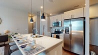 This single-floor residence is thoughtfully designed with a contemporary aesthetic, featuring consistent dark hardwood-style flooring that flows throughout the space and ties each room together cohesively. The entryway offers a welcoming introduction, complete with a brass wall-mounted hook rack for convenient storage and framed artwork that sets a stylish tone from the moment you step inside.

The open-concept kitchen is a standout feature, showcasing light wood cabinetry extending to the ceiling, a full suite of stainless steel appliances including a french-door refrigerator, electric range, and built-in microwave. A large center island with a crisp white countertop and an integrated sink serves as both a prep space and a casual dining area, illuminated by a pair of modern matte black pendant lights.

The living area benefits from multiple windows that bring in generous natural light, with views to the outdoors. The space comfortably accommodates a full seating arrangement and has a calm, airy ambiance.

The two bedrooms are well-proportioned, each featuring large windows with natural light and dark hardwood flooring. The primary bedroom includes access to a dedicated walk-in closet with wire shelving. The bathroom features tile flooring, a single-sink vanity with light wood cabinetry, and a full tub-shower combination with classic subway tile surround.