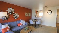 This two-floor residence offers a thoughtfully designed interior that balances bold design choices with functional living spaces. The main living area is anchored by a striking burnt-orange accent wall, which adds warmth and visual interest to the room. A decorative star-shaped mirror and a pair of coordinating art prints contribute to the curated aesthetic of the space.

The living area accommodates a comfortable gray sofa dressed with patterned and accent throw pillows, positioned around a round wooden coffee table. Neutral-toned carpeting runs throughout, softening the overall look and providing a warm underfoot feel.

Flowing seamlessly from the living area, the dining space features a compact round table with sleek metal legs, surrounded by contemporary upholstered chairs in a light gray tone. This area benefits from natural light through a nearby window, brightening the space.

Adjacent to the dining area, a pass-through or small bar area is visible, hinting at a connected kitchen beyond. Track lighting overhead provides directed illumination. The overall ambiance of the space is warm, modern, and cohesive, with a consistent color palette tying each area together.