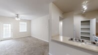 This single-floor residence offers a well-organized layout with a warm, neutral color scheme throughout. The spacious living area is carpeted in a soft tan tone and features a ceiling fan with integrated lighting, a large window that invites natural light, and an exterior door providing convenient access. Built-in open shelving along one wall adds practical display and storage space. The kitchen is positioned adjacent to the living area in a pass-through configuration, featuring warm wood-toned cabinetry, light countertops, a white electric range, a refrigerator, and a dedicated pantry closet with multiple shelves for ample storage. Dark hardwood-style flooring in the kitchen and bathroom provides a visual contrast to the carpeted areas. Bedrooms are generously sized with plush carpet, neutral walls, and closet access. The bathroom includes a full tub-shower combo with tiled surround, a single-sink vanity with matching wood-toned cabinetry, and dark hardwood-style flooring. Throughout the residence, consistent white trim, HVAC vents, and clean wall finishes contribute to a cohesive and functional interior. The floor plan flows logically between spaces, making it practical and easy to navigate.