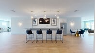 Located at 5085 Main St in Trumbull, Connecticut, this thoughtfully designed amenity space spans a single generous floor, offering a refined blend of social and functional areas finished with consistent modern aesthetics throughout.

The centerpiece of the space is a sleek bar and kitchenette area featuring a long, dark-countertop island with seating for four, flanked by white shaker cabinetry, a stainless steel refrigerator, and elegant glass pendant lighting with brass accents. A wall-mounted TV and framed artwork complete the social atmosphere.

Adjacent to the bar, a bright lounge area features light wood-look flooring in a classic herringbone pattern, a shiplap-surround fireplace with an integrated TV, and a comfortable seating arrangement. French doors open to an outdoor area, flooding the space with natural light and providing views of the surrounding community.

A sun-drenched dining nook with large windows overlooking the exterior courtyard and street offers an inviting spot for casual gatherings. The space also includes a dedicated conference room fitted with a wall-mounted display, a round table, and upholstered office chairs atop a patterned carpet — ideal for private meetings or focused work.

Wide, open corridors with recessed lighting and clean white walls connect all areas seamlessly, reinforcing the space's bright, contemporary character.
