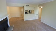 This single-floor residence features an open-concept layout that seamlessly connects the living, dining, and kitchen areas. The spacious carpeted living area is finished in neutral beige tones, creating a clean and understated ambiance throughout the space.

The kitchen is positioned centrally within the unit, accessible via a pass-through counter that opens to the main living area. It is equipped with stainless steel appliances, including a refrigerator and a gas range with an overhead exhaust hood. Light-toned cabinetry and a countertop workspace provide practical storage and preparation areas. The pass-through design enhances the sense of openness while maintaining a distinct kitchen zone.

A hallway visible toward the rear of the unit leads to additional rooms, suggesting a functional layout with separated living and private spaces. Ceiling-mounted HVAC vents are integrated throughout, ensuring climate control across the unit.

The walls are painted in a soft, warm white, complementing the beige carpeting underfoot. The overall design is practical and move-in ready, prioritizing functionality and a cohesive, neutral aesthetic suitable for a variety of furnishing styles.