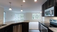 This single-floor space showcases an open-concept layout that seamlessly connects the kitchen and living area, creating a cohesive and functional environment. The kitchen is appointed with dark espresso cabinetry that provides a striking contrast against the light-toned countertops, which extend across both the perimeter surfaces and a central island. Stainless steel appliances, including a dishwasher and range, add a modern, polished touch. Pendant lighting fixtures hang above the kitchen island, complementing the recessed ceiling lights that illuminate the space evenly throughout.

The transition from the hardwood-style kitchen flooring to plush carpeting in the adjoining living area subtly delineates the two zones while maintaining an open, airy feel. The living area benefits from an abundance of natural light, courtesy of multiple large windows that frame views of mature trees and lush greenery, bringing a sense of the outdoors inside. The clean white walls and neutral color palette throughout contribute to a bright, welcoming ambiance. Crown molding adds a refined architectural detail, enhancing the overall polished aesthetic of this well-designed single-floor space.