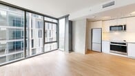 Located at 707 Terry Ave in Seattle's First Hill neighborhood, this thoughtfully designed single-floor unit offers a contemporary urban living experience. Light hardwood flooring flows throughout, creating a warm, cohesive aesthetic that complements the clean white walls and modern finishes.

The open living area is anchored by expansive floor-to-ceiling windows that flood the space with natural light and frame views of the surrounding Seattle cityscape. A concrete-accented accent wall adds an industrial touch, while the integrated kitchen features light wood cabinetry, built-in shelving, and a full-size stainless steel refrigerator.

A frosted glass sliding partition elegantly separates the sleeping area from the main living space, offering privacy without sacrificing the open, airy feel of the unit. The bedroom includes a built-in closet with sliding white panel doors, maximizing storage efficiently.

The hallway connects the living areas to the bathroom, which is finished in a crisp, minimalist style. The bathroom features a combination tub and shower surrounded by white subway tile, a wall-mounted showerhead with handheld attachment, chrome fixtures, a wall-hung toilet, and a stone-finish vanity countertop atop a clean white base. Recessed lighting and a speckled tile floor complete the polished look.