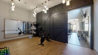Located at 9760 Rosemont Ave in Lone Tree, Colorado, this single-floor fitness center is thoughtfully designed to deliver a comprehensive workout experience. The space is divided into distinct functional zones, each with its own character and purpose. A dedicated studio area features warm hardwood-style flooring, a full-length mirror wall, and a dramatic black barn-style sliding door that serves as a bold architectural centerpiece. Pendant lighting with brass accents adds an upscale ambiance to this zone, which is equipped with a Peloton bike, stability balls, and hand weights in a range of sizes. The main strength and cardio floor transitions to durable light gray flooring and is outfitted with a variety of cable machines, multi-station strength equipment, treadmills, an elliptical, and functional fitness accessories including TRX straps and plyo boxes. Ceiling fans and large windows allow natural light to fill the space while maintaining a comfortable environment. A floor-to-ceiling steel-framed glass partition elegantly separates the fitness area from an adjoining lounge or common area, enhancing the sense of openness. The combination of modern industrial finishes, purposeful equipment layout, and refined lighting makes this fitness center both functional and visually striking.