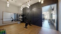 Located at 9760 Rosemont Ave in Lone Tree, Colorado, this single-floor fitness center is thoughtfully designed to deliver a comprehensive workout experience. The space is divided into distinct functional zones, each with its own character and purpose. A dedicated studio area features warm hardwood-style flooring, a full-length mirror wall, and a dramatic black barn-style sliding door that serves as a bold architectural centerpiece. Pendant lighting with brass accents adds an upscale ambiance to this zone, which is equipped with a Peloton bike, stability balls, and hand weights in a range of sizes. The main strength and cardio floor transitions to durable light gray flooring and is outfitted with a variety of cable machines, multi-station strength equipment, treadmills, an elliptical, and functional fitness accessories including TRX straps and plyo boxes. Ceiling fans and large windows allow natural light to fill the space while maintaining a comfortable environment. A floor-to-ceiling steel-framed glass partition elegantly separates the fitness area from an adjoining lounge or common area, enhancing the sense of openness. The combination of modern industrial finishes, purposeful equipment layout, and refined lighting makes this fitness center both functional and visually striking.