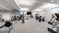 Located at 208 Woolwich St S, this fitness facility offers a thoughtfully designed, single-floor space ideal for a full-body workout experience. The room is finished with neutral grey carpet tiles and light-toned walls, creating a clean, modern aesthetic throughout. Recessed and suspended linear lighting fixtures provide bright, even illumination across the entire floor, complemented by natural light from a series of horizontal windows positioned along the lower portion of the walls.

The facility is comprehensively equipped to support a wide variety of training styles. The cardio section features treadmills, stationary bikes, and a rowing machine, while the strength training area includes a multi-station cable machine, a dumbbell rack with a weight bench, and resistance accessories. A large wall-mounted flat-screen television serves as a central focal point, supporting both guided workouts and general entertainment. Full-length mirrors along one wall enhance the sense of space and allow users to monitor their form during exercise.

Stability balls, BOSU trainers, and step platforms round out the functional training options. A dedicated entry door provides convenient access, and subtle design details such as ceiling soffits and recessed lighting zones add architectural dimension to the space. Overall, the facility presents a practical, well-maintained environment suited to diverse fitness needs.