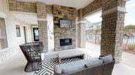 The Meadows community clubhouse patio is a well-appointed covered outdoor living space designed for year-round social gatherings and relaxation. The area is defined architecturally by prominent natural stone columns and matching stone accent walls that lend a cohesive, upscale aesthetic throughout. A floor-to-ceiling stacked stone fireplace serves as the central focal point of the lounge area, with a wall-mounted flat-screen television positioned above it, creating an ideal setting for both casual conversation and entertainment.

The lounge zone is furnished with a comfortable gray sectional sofa and a concrete-top coffee table, anchored by a bold black-and-white geometric patterned outdoor rug. Adjacent to the lounge, a distinct dining area features a concrete-style dining table paired with vibrant red metal chairs, set atop a matching geometric rug, offering a functional space for outdoor meals.

The covered patio opens on multiple sides, allowing natural light and fresh air to flow freely through the space. Direct gated access to the community swimming pool is visible from the lounge area, providing a seamless connection between amenity spaces. The entry to the adjacent interior clubhouse is marked by double glass doors and a branded welcome mat, completing a polished and welcoming community hub.