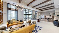Located at 3260 Stockton Street in North Fort Myers, Florida, this sophisticated community amenity space spans a single floor of impressive proportions. The grand main living area immediately commands attention with its soaring double-height ceilings, exposed wood beam accents, and floor-to-ceiling black-framed windows that flood the space with natural light and offer outdoor views of the surrounding landscape. A striking sputnik-style chandelier anchors the primary lounge, which features bold mustard-yellow seating, patterned area rugs, and a mix of modern furnishings arranged for both social gathering and relaxed conversation. An open-concept combined dining and living area flows seamlessly from the main lounge, highlighted by a long bar counter with pendant lighting, wood-look flooring, and a vibrant blue accent wall adorned with geometric mirrors. A secondary living room offers a more intimate setting, furnished with a charcoal sofa, marble coffee table, and a sculptural multi-globe chandelier. The dedicated office features a large conference table beneath a dramatic looping LED pendant light, a wall-mounted display screen, and curated artwork. A cozy private meeting nook with velvet chairs and a marble-top table completes the office suite. The hallway connects all areas with clean lines, wood-look flooring, and warm, recessed lighting throughout.