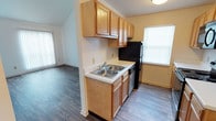 This two-story residence offers a practical and comfortable layout designed to maximize everyday functionality. The kitchen is a standout feature, equipped with warm honey-toned wood cabinetry that spans both upper and lower sections, providing ample storage space. A double-basin stainless steel sink sits at the center of the countertop workspace, complemented by light-colored laminate countertops. Black appliances, including a full-size refrigerator and dishwasher, add a sleek contrast to the warm wood tones throughout the kitchen. The kitchen flows openly into the adjacent living area, creating a connected and sociable floor plan. Durable wood-style laminate flooring runs throughout the main level, lending a cohesive and clean aesthetic to the space. Natural light enters through windows fitted with horizontal blinds, brightening the interior without sacrificing privacy. Vaulted ceiling lines are visible from the kitchen area, contributing a sense of added vertical space and architectural interest. Vertical blind window treatments in the adjoining living area further enhance the light control options available throughout the home. The two-floor design offers separation between living and private spaces, making this residence both practical and well-organized for daily living.