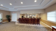 This single-floor common space is designed as a social gathering area centered around a prominent bar-style counter crafted from rich, dark wood with a dark stone or laminate surface. A row of matching dark wood bar stools lines the counter, providing ample seating for residents or guests. The counter is accented by decorative floral arrangements and framed artwork hung along the back wall, adding a warm, welcoming character to the space.

The ceiling features a stepped tray design with recessed lighting, lending a subtle architectural detail that elevates the overall aesthetic. Neutral beige and tan tones dominate the walls, complemented by a wainscoting-style accent along the lower portion. The flooring transitions from light ceramic tile near the bar area to a patterned carpet in the surrounding space, defining distinct zones within the room.

Natural and artificial lighting combine to keep the area bright and inviting. A standard door on the left provides access to adjacent spaces, suggesting this room is part of a larger facility. Overall, the space conveys a comfortable, community-oriented atmosphere well-suited for casual socializing and gatherings.