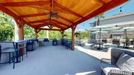This two-floor residential community, known as Riviera South, offers a thoughtfully designed outdoor amenity space that serves as a central gathering hub for residents. The centerpiece is a generously sized covered pavilion constructed with rich, warm-toned timber framing and a steeply pitched wood-plank ceiling. Dual ceiling fans keep the area comfortable year-round, while open sides allow for natural airflow and unobstructed views of the surrounding greenery.

Beneath the pavilion, a built-in grilling station is constructed from stone masonry and equipped with multiple grills, accompanied by bar-height seating ideal for casual outdoor dining. The adjacent open-air patio extends the entertaining space considerably and features multiple picnic-style tables shaded by large teal market umbrellas. Modern planters with ornamental grasses and a tall palm tree add a resort-like ambiance to the space.

A cornhole board branded with the Riviera South logo adds a recreational element to the patio, reinforcing the community-oriented character of the property. The surrounding grounds are well-maintained with manicured lawns, mature trees, and a children's playground visible in the background. The two-floor residential buildings feature a classic combination of siding and brick exterior, complementing the welcoming and vibrant outdoor environment.