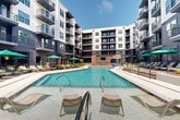 Located at 4720 Nations Crossing Rd in Charlotte, NC, this outdoor amenity courtyard is a standout feature of a modern multi-story residential community. The space is defined by a large, resort-style swimming pool with crystal-clear turquoise water, flanked on multiple sides by expansive concrete pool decks lined with lounge chairs and green shade umbrellas, ideal for sunbathing and relaxation.

The courtyard is thoughtfully designed with distinct zones that cater to a variety of outdoor activities. A raised entertainment wall features a mounted flat-screen TV beneath a pergola-style overhang, creating a dedicated outdoor viewing area directly adjacent to the pool. Stepped terraces with raised planting beds and ornamental landscaping provide natural transitions between levels, while string lights draped overhead lend an inviting ambiance for evening gatherings.

A lush, well-manicured artificial turf lawn area offers additional recreational space, complemented by Adirondack-style seating and curated landscaping featuring boulders, shrubs, and young trees. The entire courtyard is enclosed by the surrounding multi-story building, finished in a modern palette of deep charcoal, white, and dark trim, with private balconies overlooking the amenity space below. The design balances functionality with a sophisticated, resort-inspired aesthetic.
