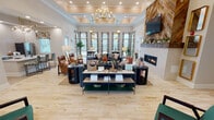 Located at 1000 Infinity Drive in Maryville, Tennessee, this single-floor community clubhouse offers a comprehensive range of amenity spaces designed for comfort, productivity, and wellness. The centerpiece is a generous open lounge area distinguished by soaring coffered ceilings, an elegant multi-arm chandelier, and abundant natural light streaming through large, curtain-framed windows. Plush seating arrangements surround a striking fireplace flanked by a herringbone wood-plank accent wall with a mounted television, creating a warm and inviting gathering space. A full kitchenette with modern appliances is seamlessly integrated nearby, alongside bar-height seating for casual dining. The fitness center is expansively equipped with treadmills, ellipticals, rowing machines, weight racks, and strength-training machines, all set beneath recessed lighting with natural light from multiple windows. A dedicated business center provides workstation computers and comfortable lounge seating, while a separate room offers a textured accent wall, worktable, and built-in cabinetry for creative or multi-purpose use. A formal conference room features a large table beneath a statement wood-and-metal chandelier with tall ceilings. Connecting hallways are adorned with original artwork, tile flooring, and dual water stations, reinforcing the thoughtful and polished design throughout.