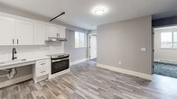Located at 101 Cedar St in Providence, Rhode Island, this single-floor unit offers a thoughtfully updated interior with a clean, contemporary aesthetic. The kitchen is a standout feature, outfitted with white shaker-style cabinetry, open shelving, stainless steel appliances including a refrigerator, dishwasher, and range with hood, and a utility sink — all set atop light wood-look luxury vinyl plank flooring that extends through the main living areas. The layout, as revealed in the dollhouse view, presents a well-organized floor plan with distinct living, sleeping, and utility zones connected by clear corridors. The bedrooms are carpeted with a patterned blue-gray tile-style carpet and feature neutral gray walls, white baseboard heating, and windows that frame elevated views of the Providence cityscape, filling the rooms with natural light. One bedroom includes a sliding-door closet with built-in hanging rod, adding practical storage. The bathroom is finished with the same wood-look flooring, a floating white vanity with a large mirror, and clean white fixtures, offering a bright and accessible design. Throughout the unit, details such as black hardware accents, white trim, and a neutral color palette create a cohesive and modern living environment.