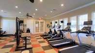 This single-floor fitness center is thoughtfully designed to provide a comfortable and functional workout environment. The space is outfitted with a row of modern cardio equipment, including treadmills and elliptical machines, each equipped with individual display screens for an enhanced exercise experience. A freestanding adjustable seat adds versatility for strength or stretching exercises.

The flooring features a bold, checkered pattern in shades of orange, brown, and beige, adding a vibrant visual energy to the space. Recessed lighting throughout the ceiling ensures bright, even illumination, while two ceiling fans promote air circulation and comfort during workouts. Crown molding along the ceiling adds a refined architectural touch.

Natural light flows in generously through multiple windows fitted with white blinds, offering views of the surrounding greenery and creating an open, airy atmosphere. Elegant French doors with glass panels serve as a stylish entryway into the fitness area, enhancing the overall aesthetic. The warm, neutral wall tones complement the lively flooring and contribute to an inviting ambiance. Overall, this fitness center balances practicality with thoughtful design, making it a welcoming space for daily exercise.
