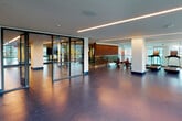 This contemporary, two-floor fitness center is defined by its impressive architectural design and extensive range of workout spaces. Towering floor-to-ceiling glass curtain walls flood the main cardio floor with abundant natural light while offering wide panoramic views of the surrounding outdoor landscape, including tree-lined streetscapes and neighboring buildings.

The primary cardio area accommodates a long row of treadmills, elliptical trainers, stationary bikes, and rowing machines, all positioned to take full advantage of the exterior views. Durable, speckled blue rubber flooring runs throughout, providing both a clean aesthetic and a functional, high-performance surface.

A dedicated strength and free weights zone features a well-curated selection of dumbbells, benches, and cable machines. This area is distinguished by warm wood slat accent walls backlit with ambient lighting, creating a bold visual contrast to the otherwise clean, minimalist palette. Full-length mirrors amplify the sense of space and assist with form during training.

The upper floor offers additional workout areas, including space for cycling equipment and functional training. Glass railings overlook the lower level, maintaining visual connectivity between floors. Linear LED lighting provides consistent, modern illumination throughout both levels, complementing the overall sleek and professional atmosphere of the facility.