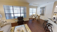 This well-appointed single-floor residence in Dallas offers a seamless blend of classic detailing and modern comfort. The open-concept layout connects the living and dining areas, creating a spacious and inviting atmosphere ideal for both everyday living and entertaining.

Rich, dark hardwood flooring flows throughout the main living spaces, adding warmth and a sense of elegance. Crown molding frames the ceilings, lending a refined architectural touch that complements the neutral, creamy wall tones. Large windows fitted with horizontal blinds allow abundant natural light to pour into the space while providing views of the surrounding greenery outdoors.

The living area is anchored by a glass-top coffee table and features a sleek accent chair, offering a comfortable and contemporary setting. A bold yellow geometric area rug introduces a pop of color and visual interest. The adjacent dining area is furnished with a round glass-top table surrounded by upholstered chairs, accompanied by modern track lighting overhead and striking wall art that reinforces the space's sophisticated aesthetic.

Overall, the residence exudes a bright, clean, and polished ambiance, with thoughtful design details throughout that create a cohesive and comfortable living environment in the heart of Dallas.