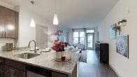 This thoughtfully designed single-floor residence offers a seamless blend of modern style and functional living. The open-concept kitchen is a standout feature, showcasing rich dark cabinetry, stainless steel appliances, a white subway tile backsplash, granite countertops, and a central island with pendant lighting overhead — all flowing effortlessly into the living area. The living room is bright and welcoming, with large windows dressed in sheer curtains that invite natural light and provide views of the surrounding exterior. Wide-plank hardwood-style flooring runs throughout the main living areas, adding warmth and continuity to the space. The residence includes multiple bedrooms, each carpeted for comfort and featuring generous windows. One bedroom connects directly to a private en-suite bathroom and a spacious walk-in closet with built-in shelving and hanging rods. The bathrooms are elegantly appointed with granite-topped dark wood vanities, large frameless mirrors, and tile flooring. A full bathroom with a soaking tub and a separate full-vanity bathroom serve the additional bedrooms. The overall ambiance is clean, contemporary, and welcoming, with a neutral color palette that creates a calm, cohesive atmosphere throughout.