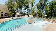 Located at 1421 Legacy Falls Dr in Wake Forest, NC, this beautifully designed outdoor amenity area offers a resort-inspired experience within a lush, tree-lined setting. The centerpiece is a generously sized freeform pool featuring a stone-inlaid sun shelf with in-water lounge chairs, multiple entry points, and a secondary wading area. The pool deck is outfitted with cabana-style canopies, lounge chairs, and shade umbrellas, creating an inviting retreat ideal for relaxation. Adjacent to the pool, a flagstone patio surrounds a glass-crystal fire pit, furnished with contemporary upholstered seating and a large market umbrella—perfect for outdoor gatherings. A striking stacked-stone accent wall anchors a more intimate seating nook, adding a rugged yet refined architectural element to the space. The building exterior showcases classic shake-style siding in a soft neutral palette, complemented by large white-trimmed windows and a herringbone-patterned stone tile walkway that adds refined detail underfoot. Wrought iron fencing and gated entries provide security and definition throughout the outdoor areas, while elevated vantage points offer views of the pool and surrounding mature tree canopy. The overall ambiance is polished, tranquil, and resort-like.