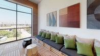 This contemporary rooftop lounge is a thoughtfully designed communal space that blends indoor comfort with an open, airy atmosphere. The space is unified by warm teak-style interlocking deck flooring that runs throughout, providing a cohesive and inviting foundation underfoot.

Floor-to-ceiling windows and glass doors line the perimeter, flooding the interior with abundant natural light and offering sweeping panoramic views of the surrounding cityscape and skyline beyond. The expansive glazing creates a seamless visual connection between interior and exterior, making the urban surroundings a defining feature of the space.

The lounge is organized into distinct seating areas suited for both social gatherings and quieter moments. A built-in banquette with a solid wood pallet-style base is dressed in deep navy cushions and vibrant green accent pillows, positioned beneath a pair of bold, colorful canvases that add artistic character to the white walls. A compact green sofa and light wood side tables create an intimate conversation nook nearby, while a dining-height table with woven chairs offers a more structured setting.

A dramatic floor-to-ceiling grey tile accent wall anchors the central zone, complemented by a wall-mounted display screen. Warm globe wall sconces and recessed ceiling lighting provide a layered, ambient glow. Built-in cabinetry with a warm wood finish adds functional storage, completing this well-appointed and versatile lounge environment.