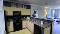 This single-floor space showcases a thoughtfully designed kitchen as its centerpiece, finished with a striking combination of navy blue accent walls and crisp white crown molding that adds a polished architectural detail. The kitchen is outfitted with cream-toned cabinetry, offering generous upper and lower storage space, complemented by dark countertops that provide a practical and stylish work surface. Black appliances — including an electric range, over-the-range microwave, dishwasher, and refrigerator — create a cohesive, contemporary look throughout the cooking space. A double-basin stainless steel sink is conveniently positioned along the countertop run, with ample cabinet storage below. Decorative accents above the upper cabinets, including framed artwork and sculptural pieces, add personality and visual interest to the upper wall space. A charming arched pass-through window opens from the kitchen into the adjacent living area, enhancing the sense of connectivity and allowing natural light to flow between spaces. The light tile flooring ties the kitchen together with a clean, neutral foundation. Overall, the space presents a functional yet characterful interior with a bold color palette and well-considered design details.
