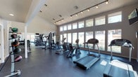 This single-story fitness center offers a thoughtfully designed workout environment that balances functionality with an open, airy atmosphere. The space features soaring ceilings that enhance the sense of spaciousness, while a row of large, floor-to-ceiling windows along one wall floods the interior with abundant natural light and provides pleasant views of the surrounding outdoor greenery. The flooring consists of durable, dark rubberized material well-suited for high-traffic exercise use. Modern track lighting runs across the ceiling, supplementing natural light and ensuring the space remains bright throughout the day. The facility is equipped with a variety of cardio machines, including treadmills and elliptical trainers, positioned to take advantage of the window views. Toward the rear of the space, a selection of strength and cable training machines is arranged alongside a large wall-mounted mirror, which visually expands the room and allows for form monitoring during workouts. The overall ambiance is clean, modern, and welcoming, creating an environment well-suited for a productive fitness routine.