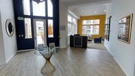 This single-floor community space serves as a polished and professional welcome center, designed to make an immediate impression on visitors. The layout is open and airy, anchored by dramatically tall arched windows flanking the main entrance that flood the interior with abundant natural light. The entryway features light-toned hardwood-style flooring that flows seamlessly throughout the space, contributing to a clean and contemporary aesthetic.

Near the entrance, a sculptural glass-top accent table with a woven base provides an elegant focal point and a place for informational displays. The reception desk, finished in dark wood tones, is positioned centrally within the space and set against a striking bold yellow feature wall, which adds energy and visual warmth to the room. Surrounding windows on multiple sides offer views of the surrounding residential community, reinforcing a sense of connection to the neighborhood.

Recessed lighting complements the natural light, ensuring the space remains bright and inviting throughout the day. Seating areas with upholstered blue chairs are visible in the background, offering comfortable spaces for consultations or casual waiting. The overall ambiance is modern, organized, and welcoming — reflecting a thoughtfully designed environment intended to inform and engage visitors effectively.