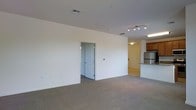 This single-floor space offers an open and functional layout designed to maximize usability. The main living area is generously proportioned, featuring neutral-toned carpeting and white walls that create a bright, clean atmosphere. A track lighting fixture is mounted on the ceiling, providing focused illumination across the space, complemented by a ceiling vent that ensures consistent climate control.

An open doorway connects the living area to an adjacent room, likely a bedroom or utility space, maintaining a sense of flow throughout the unit. Toward the rear, the layout transitions into a kitchen area, distinguished by warm wood cabinetry, a granite or stone-topped breakfast bar, and what appears to be stainless steel appliances. The kitchen benefits from its own ceiling light fixture, keeping the area well-lit and practical.

The overall design emphasizes simplicity and versatility, with neutral finishes that lend themselves to a variety of furnishing styles. The open-plan arrangement between the living and kitchen areas encourages easy interaction and everyday functionality, making the space well-suited for comfortable day-to-day living.