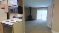 This single-floor space offers a practical and comfortable layout that combines a functional kitchen with an open living area. The kitchen is well-equipped, featuring a coil-burner electric range, an over-the-range microwave, and wood-toned cabinetry that provides ample storage. A half-wall partition separates the kitchen from the main living area, creating a sense of division while maintaining an open feel. The living area is carpeted throughout and centers around a standout freestanding black wood-burning fireplace suspended from the ceiling — a distinctive architectural focal point that adds warmth and character to the room. Large sliding glass doors dressed with vertical blinds span one wall of the living area, allowing generous natural light to flood the space and offering views of the surrounding greenery outdoors. The neutral tones of the walls and flooring lend the space a calm, understated ambiance, while the fireplace provides a bold contrast. The overall layout is efficient and well-considered, making the most of the available space on a single floor.