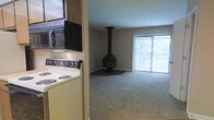 This single-floor space offers a practical and comfortable layout that combines a functional kitchen with an open living area. The kitchen is well-equipped, featuring a coil-burner electric range, an over-the-range microwave, and wood-toned cabinetry that provides ample storage. A half-wall partition separates the kitchen from the main living area, creating a sense of division while maintaining an open feel. The living area is carpeted throughout and centers around a standout freestanding black wood-burning fireplace suspended from the ceiling — a distinctive architectural focal point that adds warmth and character to the room. Large sliding glass doors dressed with vertical blinds span one wall of the living area, allowing generous natural light to flood the space and offering views of the surrounding greenery outdoors. The neutral tones of the walls and flooring lend the space a calm, understated ambiance, while the fireplace provides a bold contrast. The overall layout is efficient and well-considered, making the most of the available space on a single floor.