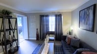 This single-floor space showcases a thoughtfully designed living room that serves as its centerpiece. The room is finished with neutral gray walls accented by crisp white crown molding, lending a clean and polished aesthetic throughout. Dark hardwood-style flooring grounds the space and adds warmth, complementing the overall color palette.

A large window dressed with charcoal grommet-panel curtains allows generous natural light to filter into the room, creating a bright and welcoming atmosphere. The window treatment adds a contemporary touch while offering privacy and light control. Decorative framed wall art adds a subtle artistic element to the space.

The living area is furnished with a navy blue sofa adorned with accent pillows in warm earth tones, paired with a rectangular coffee table that anchors the seating arrangement. A patterned area rug defines the lounge area. Additional decor elements, including a tall decorative vase and a floor lamp with a plant, contribute texture and visual interest. A doorway to the left hints at access to additional areas or an outdoor space, suggesting a well-connected and functional layout throughout the home.