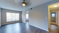 This single-floor space offers a clean, contemporary layout designed for comfortable everyday living. The main living area is open and airy, showcasing rich hardwood-style flooring that extends seamlessly throughout the space, creating a warm yet modern aesthetic. Neutral gray walls provide a versatile backdrop that complements a wide range of furnishing styles.

Natural light is a defining feature of this space, with multiple windows fitted with white horizontal blinds allowing residents to control brightness and privacy with ease. A ceiling fan with an integrated light fixture keeps the living area well-lit and comfortable year-round.

A visible passageway connects the main living area to an adjacent room, which also benefits from its own window and natural light, suggesting a well-connected and functional floor plan. The textured ceiling adds a subtle architectural character to the interior.

Thoughtfully placed electrical outlets, HVAC vents, and clean white baseboards reflect a well-maintained interior. The overall ambiance is bright, neutral, and move-in ready, offering a welcoming environment that is both practical and visually appealing.
