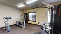 This single-level fitness center is designed to accommodate a comprehensive workout experience, offering a thoughtfully arranged selection of exercise equipment across a spacious, open floor plan. The room features neutral cream-colored walls accented by dark brown baseboards and trim, creating a clean and functional aesthetic throughout. Textured ceilings with flush fluorescent lighting panels provide consistent, bright illumination across the space.

The cardio section is well-stocked, housing a treadmill, elliptical trainer, and stationary bike, each positioned to allow ample room for comfortable use. A dedicated strength-training area features a Life Fitness multi-station cable machine, adjustable weight benches, and free weights, catering to a range of resistance-based exercises. Full-length wall mirrors line one section of the room, visually expanding the space while allowing users to monitor their form during workouts.

Natural light filters in through several windows fitted with horizontal blinds, adding brightness to the carpeted interior. The space also includes a wall-mounted hand sanitizer station near the entry door and clearly marked emergency exit signage, reflecting attention to safety and hygiene. The overall ambiance is practical and well-maintained, offering a welcoming environment for fitness-focused use.