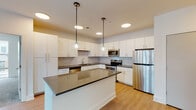 This contemporary single-floor residence showcases a clean, modern design with consistent finishes throughout. The heart of the home is a well-equipped kitchen featuring crisp white cabinetry, dark countertops, and a classic subway tile backsplash. A central island with pendant lighting provides additional prep space and a natural gathering point. Stainless steel appliances — including a refrigerator, range, microwave, and dishwasher — complete the functional and stylish setup. Light wood-tone flooring runs through the kitchen and main living areas, adding warmth to the bright, neutral palette. The open living area benefits from natural light streaming through large windows and sliding glass doors, with a ceiling fan overhead for year-round comfort. Bedrooms are carpeted and generously sized, each featuring ceiling fans and expansive windows that frame elevated urban views of the surrounding cityscape. The primary bathroom offers a wide vanity with dark countertops, white cabinetry, wood-look flooring, and access to a walk-in closet with built-in shelving. A second bathroom includes a tub-shower combination with vertically laid white tile, a matching vanity, and the same cohesive flooring. Throughout the residence, the design maintains a consistent modern aesthetic with clean lines, neutral tones, and quality fixtures.