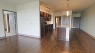 This single-floor residence offers a thoughtfully designed open-concept layout that seamlessly connects the kitchen and living areas. Rich, dark hardwood flooring flows throughout, lending warmth and continuity to the space.

The kitchen is a standout feature, appointed with dark espresso cabinetry, light granite countertops, and a full suite of stainless steel appliances, including a refrigerator, microwave, and range. A kitchen island with a built-in sink and elegant pendant lighting above serves as both a functional workspace and a natural gathering point. Track lighting along the ceiling provides additional illumination across the cooking area.

The open living area is generously proportioned, offering flexible space for a variety of furniture arrangements. Neutral wall tones create a clean, bright backdrop that complements the darker flooring and cabinetry. A hallway visible from the main living area leads to additional rooms, suggesting a practical and well-organized floor plan. The overall ambiance is modern and polished, combining contemporary finishes with a warm, inviting atmosphere suited for everyday living.