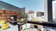 This contemporary rooftop terrace is a thoughtfully designed outdoor retreat situated above a vibrant urban landscape. The space is defined by a clean, modern aesthetic that blends natural warmth with sleek architectural detail. Horizontal warm-toned wood slat screens and pergola structures create partial enclosure and visual interest, offering both shade and privacy while maintaining an open, airy feel.

The terrace is organized into distinct seating zones. One area features a generous sectional sofa with colorful geometric throw pillows, positioned alongside large cylindrical planters filled with mature trees and lush greenery, adding a natural, garden-like quality to the rooftop. Another more intimate seating nook showcases bold yellow accent chairs and a low coffee table, set against rich wood panel walls that bring warmth and texture to the outdoor environment.

A smaller balcony area provides a quiet perch with direct, unobstructed views of the surrounding city skyline, where high-rise office towers and neighboring buildings frame the horizon. The smooth concrete flooring throughout the terrace ties the zones together cohesively.

The overall ambiance is modern and inviting, with a strong emphasis on outdoor living. The combination of curated greenery, contemporary furnishings, vibrant accent colors, and expansive cityscape views makes this rooftop terrace a standout urban amenity.