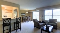 This single-floor residence features an open-concept design that seamlessly connects the living, dining, and kitchen areas into one cohesive space. The layout is anchored by plush wall-to-wall carpeting in a warm neutral tone, complemented by taupe-painted walls that create a welcoming and understated atmosphere throughout.

The dining area is defined by a classic pendant light fixture hanging above a dark wood dining table surrounded by matching chairs. A framed piece of wall art adds a decorative touch nearby. Adjacent to the dining space, a pass-through opening connects to the kitchen, where bar-height seating provides a casual spot for quick meals or conversation. The kitchen area is illuminated by its own warm lighting, offering a bright and functional workspace.

The living area is furnished with patterned upholstered armchairs and a side table, positioned to take advantage of the abundant natural light streaming in through multiple large windows. The windows, framed simply to maximize light, offer views to the outside and lend the space an airy, open feel. A textured ceiling adds subtle depth to the overall design. The single-floor layout ensures easy accessibility and a smooth flow throughout the entire space.