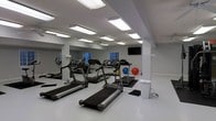 This single-floor fitness space is designed to support a practical and efficient workout experience. The room features a light gray painted interior with a smooth, easy-to-clean floor surface that contributes to a clean, clinical aesthetic. Multiple treadmills are arranged in a row as the focal point of the space, complemented by an elliptical/stationary bike machine, making cardiovascular exercise the primary focus of the facility. A large wall mirror spans one side of the room, visually expanding the space and allowing users to monitor their form during exercise. A wall-mounted flat-screen television is positioned for easy viewing from the cardio equipment. The ceiling is fitted with several rectangular fluorescent light fixtures that provide bright, even illumination throughout the room. A window, visible in the mirror's reflection, allows some natural light to enter the space. Stability and exercise balls are stored along one wall, adding versatility to the available workout options. The overall ambiance is functional and no-frills, prioritizing utility and cleanliness for fitness use.