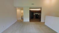 This single-floor residence offers an open and functional layout designed for practical everyday living. The main living area is finished with neutral-toned carpeting, creating a clean and understated foundation that extends throughout the space. High ceilings add a sense of openness and volume to the room, enhancing the overall airiness of the interior.

The kitchen is positioned adjacent to the living area and is separated by a half-wall, allowing for a connected yet defined space. It features dark wood cabinetry that provides ample storage, dark-toned countertops, and black appliances including a refrigerator and range with overhead ventilation. The kitchen floor transitions to a hard surface, distinguishing it from the carpeted living area.

A dedicated laundry area is visible through a separate doorway off the main living space, housing side-by-side washer and dryer units, adding in-unit convenience. The overall color palette of the home is light and neutral, with white walls and standard overhead lighting, contributing to a bright and cohesive atmosphere throughout the residence.